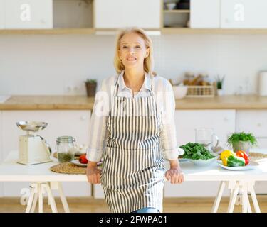 Porträt einer schönen, positiv reifen Frau in der Schürze in der Küche, die die Kamera ansah und lächelte, einer pensionierten Großmutter, die neben dem Tisch mit gesunden Zutaten stand, während sie für die Familie zu Hause kochte Stockfoto