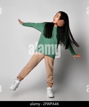 Asiatisches Mädchen in grüner Sportswear und weißen Sneakers steht auf einem Bein mit ausgestreckten Armen, balancierend Stockfoto