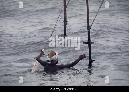 Alter Stelzenfischer mit seiner Angelrute und leerem Beutel in einer Hand und schwimmt, greifen in Richtung seiner Bambusstange in der Mirissa Strand Küste, fleißige tr Stockfoto