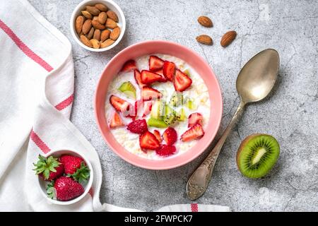 Haferflockenbrei mit Kiwi-Scheiben, Erdbeeren, Mandeln in rosa Schale, Löffel, Serviette mit roten Streifen auf Betongrund Stockfoto