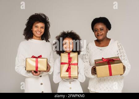 Lächelnde afroamerikanische Mutter, Tochter und Großmutter halten Geschenkboxen auf grau isoliert Stockfoto