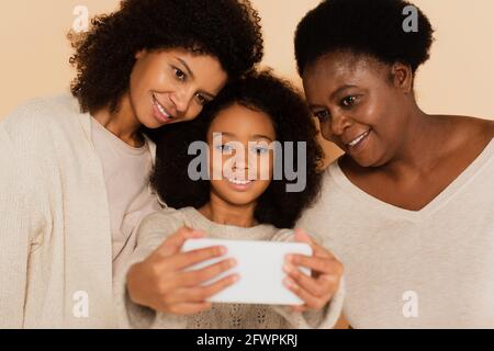 afroamerikanische Tochter, Enkelin und Großmutter, die zusammen auf beigem Hintergrund Selfie auf dem Handy machen Stockfoto