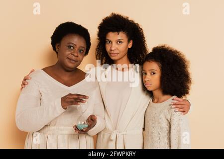 Seriöse afroamerikanische Großmutter hält Sandglas mit Tochter und Enkelin Steht in der Nähe auf beigem Hintergrund Stockfoto
