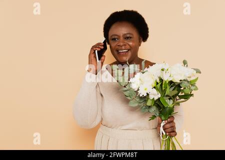 afroamerikanische Dame mittleren Alters spricht auf dem Smartphone in und Hält Blumenstrauß in der Hand auf beigem Hintergrund Stockfoto