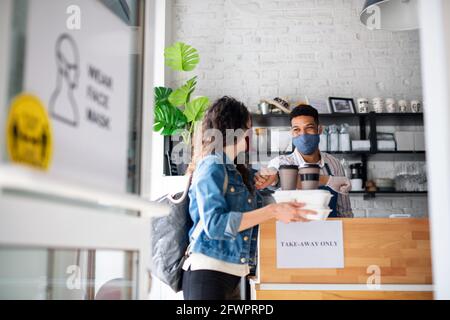 Frau mit abgepackten Speisen und Kaffee zum Mitnehmen, Begrüßung des Kellers im Café. Coronavirus-Konzept. Stockfoto