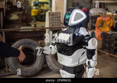 Humanoider Roboter auf Rädern mit einem Monitor auf der Brust, der einem Mann die Hände schüttelt. Die alte überladen Garage. Stockfoto