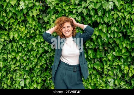 Junge Geschäftsfrau mit formeller Kleidung und lockigen Haaren lächelnd und Blick auf die Kamera vor grünem Hintergrund mit Pflanzen. Stockfoto