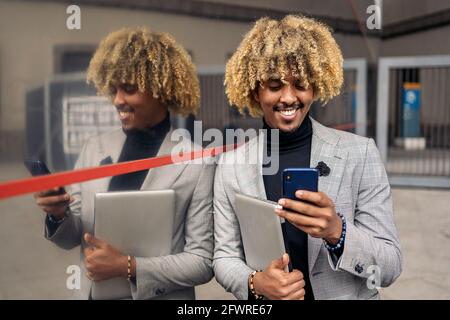 Glücklicher schwarzer Geschäftsmann mit afro Haaren und coolem Anzug, der lächelt und sein Telefon im Freien benutzt. Stockfoto