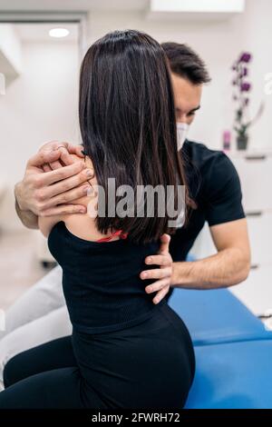 Männlicher Physiotherapeut, der Gesichtsmaske trägt und mit einem nicht erkannten Patienten in der Physiotherapieklinik arbeitet. Stockfoto