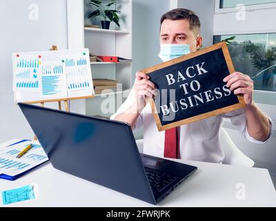 Ein Geschäftsmann in einer Maske zeigt während der Online-Kommunikation ein Zeichen zurück zum Geschäft. Stockfoto