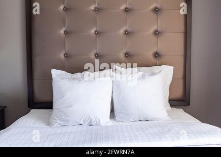 Ein makellos sauberes, fertig gemachtes Queen-Size-Bett mit weißen Laken und vier Kissenbezügen mit gepolstertem Kopfteil ist an der Wand. Ein pastellfarbenes helles aufgeräumtes c Stockfoto