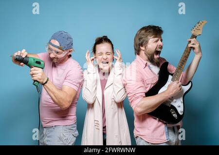 Frau in Stress bedeckt ihre Ohren, zwei Männer ärgern sie, einer spielt Gitarre, der andere arbeitet mit einem Bohrer. Ein lauter Nachbar. Stockfoto