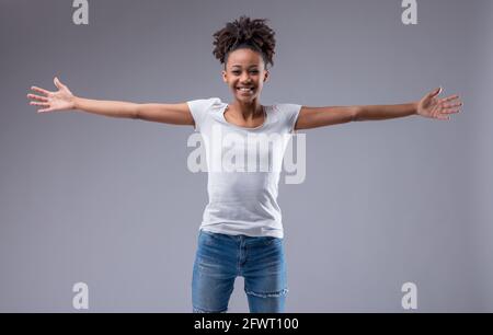 Fröhlich überschwänglich junge schwarze Frau grinst glücklich an die Kamera Als sie mit ausgestreckten Armen in Jeans und T-Shirt posiert Auf grauem Studiohintergrund Stockfoto