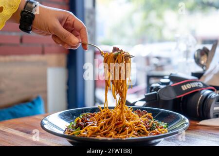 Frau mit Gabel, die die indo-chinesische mandschurische hakka-Straße abholt Nudeln aus einer schwarzen Schüssel, die auf eine Holzschale gelegt wird Tisch in einem kleinen Straßenrestaurant Stockfoto