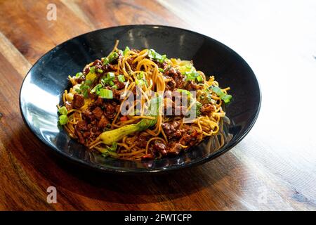 Indo chinesische dünne Nudeln garniert mit grünem Gemüse und Gewürzen Mit Chiceken Tofu und Paneer auf eine schwarze Schale gelegt Auf einem Holztisch Stockfoto