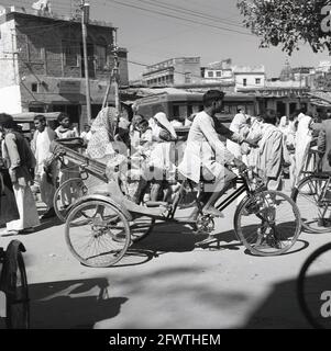 In den 1950er Jahren, ein Fahrradtaxi, Indien, eine Mutter und mehrere Kinder, die eine Fahrt im hinteren Teil der Fahrradrikscha machen, die von einem männlichen Fahrer barfuß durch eine staubige Straße gefahren wird. Dieses kleine Nahverkehrsunternehmen, eine Art Dreirad mit Heckklappe, würde die Passagiere auf Mietbasis befördern und war daher unter verschiedenen Namen bekannt: Fahrradtaxi, Velo-Taxi, Trishaw oder Hatchback-Fahrrad. Cycle Rikschas entstanden in den 1880er Jahren. In den späten 1920er Jahren waren sie in Singapur weit verbreitet und bis 1950 überall in Süd- und Ostindien zu finden. Es gibt verschiedene Designs, in Indien sitzen die Passagiere hinter dem Fahrer. Stockfoto