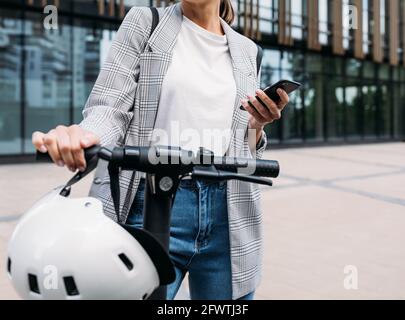 Nicht erkennbare Geschäftsfrau, die vor einem Bürogebäude mit steht Elektroroller und halten ein Smartphone Stockfoto