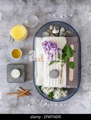 Vertikale, medizinische Zusammensetzung mit Massagezubehör, Handtüchern, Ölen und einem lila Zweig auf grauem Hintergrund. Wellness, Spa, Relax und Me Time Co Stockfoto