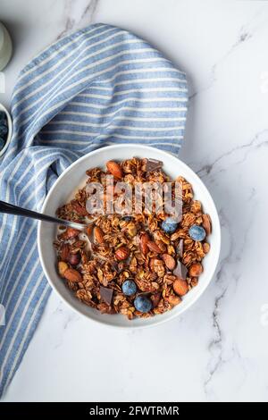 Hausgemachtes Schokoladengranola, Müsli mit Mandeln, Haselnüssen und Heidelbeeren in Schale auf weißem Marmorboden. Gesundes Frühstück. Draufsicht. Flache Lay m Stockfoto