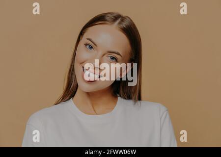 Verspielte dunkelhaarige Frau kippt den Kopf zeigt Zunge und lächelt positiv hat Spaß innen gekleidet in weißen Pullover Posen vor beigem Hintergrund. Mensch Stockfoto