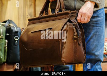 Die Hand eines Mannes, der eine braune Reisetasche vor dem Hintergrund anderer Taschen im Laden hält. Das Konzept der Naturlederprodukte Stockfoto