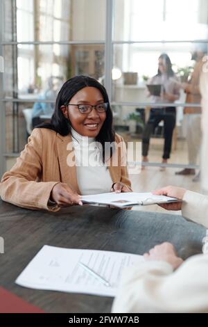 Lächelnde junge afroamerikanische Frau in Gläsern, die am Tisch und sitzen Geben ihres Lebenslaufs an die HR-Führungskraft während des Vorstellungsgesprächs Stockfoto
