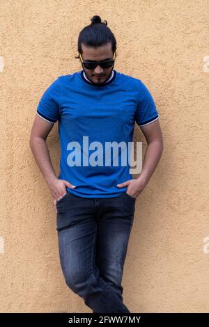 Junger Latino-Mann mit Sonnenbrille und Händen in Taschen, der auf einer senffarbenen Wand steht und nach unten blickt. Frisches Konzept Stockfoto