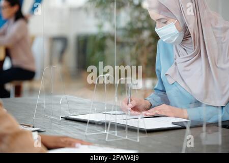 Junge arabische Frau in Hijab und Gesichtsmaske sitzend Tabelle mit Sperrsiebe und Ausfüllen von Formularen Stockfoto