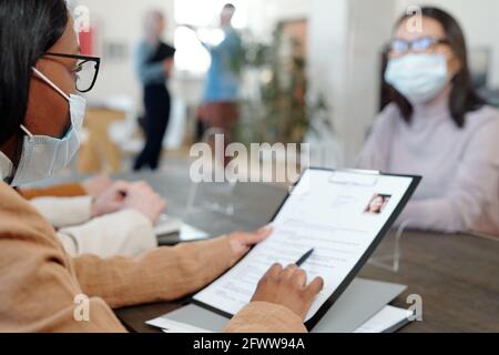 Überschulter-Ansicht des schwarzen Recruiters in Brille und Maske Sitzen am Tisch und Fragen über Berufserfahrung zu stellen Kandidat während des Interviews Stockfoto