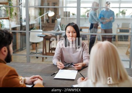 Seriöse junge asiatische Stellenbewerber sitzen am Tisch und halten Brillen, während Sie sich die Frage der Rekrutierungspersonal anhören Stockfoto