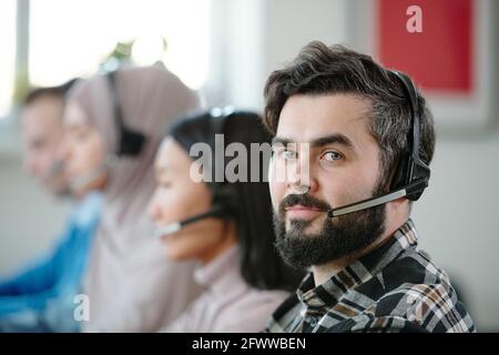 Content-Portrait junger bärtiger Telefonhörer mit schnurlosem Headset Mit Mikrofon, das gegen multiethnische Kollegen sitzt Stockfoto