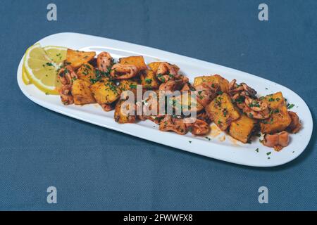 Aperitif oder typisch spanische Tapa, Tintenfisch mit Knoblauch und Kartoffeln, serviert auf einem länglichen weißen Tablett mit Zitrone und rustikalem Hintergrund. Traditionell Stockfoto