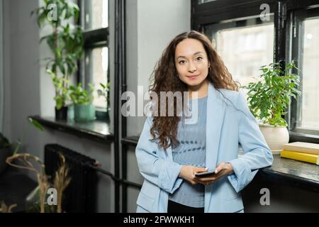 Junge Brünette Geschäftsfrau in blauem Anzug, der am Fenster steht Büro Stockfoto