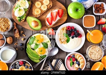 Gesunde Frühstückstisch-Szene mit Obst, Joghurt, Smoothie-Schüssel, Haferflocken, nahrhaftem Toast und Eierpfanne. Blick über einen Holzhintergrund. Stockfoto