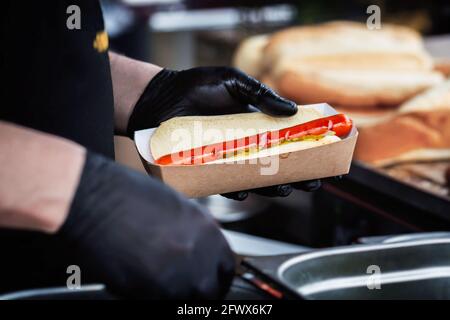 Hände des Küchenchefs, der frischen Hot Dog mit Gemüse und Sauce zubereitet. Gegrillte Wurst im Brötchen. Fast Food, Grill, Grill-Konzept Stockfoto