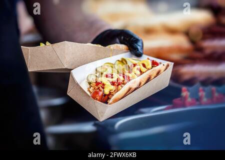 Männliche Hand des Küchenchefs hält eine Pappschachtel mit frisch zubereitetem Hot Dog mit verschiedenen leckeren Ergänzungen aus der Nähe. Grill, Grillkonzept Stockfoto