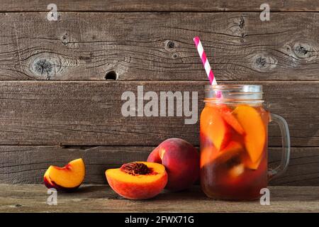 Hausgemachter Pfirsich-Eistee in einem Einmachglas-Glas. Seitenansicht mit geschnittenem Obst vor rustikalem Holzhintergrund. Stockfoto