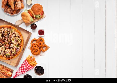 Verschiedene Speisen zum Mitnehmen oder zur Lieferung. Seitenrand. Pizza, Hamburger, gebratenes Huhn und Beilagen. Oben auf einem weißen Holzhintergrund mit Kopierraum. Stockfoto