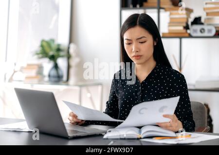 Konzentrierte seriöse attraktive erfolgreiche junge asiatische Geschäftsfrau, Managerin oder Freiberuflerin, die an einem Schreibtisch im Büro sitzt, Laptop für die Arbeit benutzt, Dokumente und Diagramme durchschaut, Bericht analysiert Stockfoto