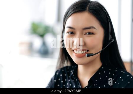 Nahaufnahme eines schönen selbstbewussten langhaarigen jungen asiatischen Frau im Headset, Freiberufler, Callcenter-Mitarbeiter oder Berater, trägt ein schwarzes stylisches Hemd, sieht direkt auf die Kamera und lächelt freundlich Stockfoto