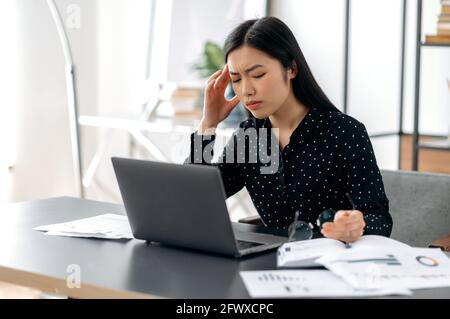 Überfordert verärgert asiatische Geschäftsfrau, Freiberuflerin oder Managerin, arbeitet am Laptop, sitzt an ihrem Arbeitsplatz, Pause, erlebt Stress, hält Brille in der Hand, schließt ihre Augen Stockfoto