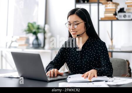 Glücklich attraktive smart freundliche junge asiatische Frau trägt Brille und Headset, Call-Center-Mitarbeiter, Experte, sitzt an einem Tisch im Büro, im Gespräch mit Kunden oder Mitarbeiter, führt Online-Beratung, Lächeln Stockfoto