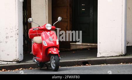 Ein süßes, leuchtend rotes Vespa-Motorroller oder Moped-Motorrad, das auf einem sanften Hügel mit einer rustikalen, grungigen Innenstadtumgebung hinter sich geparkt ist. Stockfoto