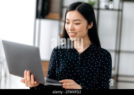 Porträt fröhlich smart niedlich erfolgreiche asiatische junge Geschäftsfrau, Manager oder Freiberufler, steht vor dem Hintergrund der verschwommenen Büro, in stilvoller Kleidung, hält offenen Laptop in den Händen, schaut Nachrichten Stockfoto