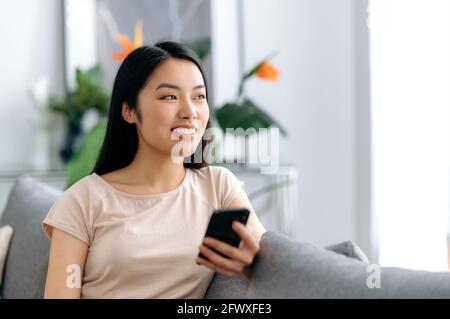 Smartphone verwenden, online chatten. Glückliches, asiatisches, hübsches Mädchen, das ihr Handy benutzt, schaut nachdenklich weg und lächelt, sitzt auf einer Couch und surft im Internet und in den sozialen Medien, schreibt mit Freunden Stockfoto