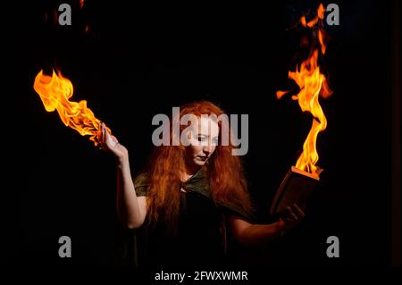 Eine ominöse Hexe setzt ihr Zauberbuch mit der Kraft des Denkens in Brand. Rothaarige Frau zaubert für Halloween. Flammen auf den Händen. Stockfoto