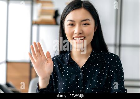 Nahaufnahme Webcam-Porträt einer freundlichen, ziemlich intelligenten, selbstbewussten jungen asiatischen Frau, Freiberuflerin oder Managerin, sitzt am Schreibtisch, plaudert mit Kollegen oder Freunden via Video-Link, winkt Hand, lächelt Stockfoto