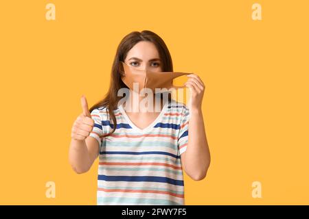 Junge Frau mit medizinischer Maske und Daumen-up auf Farbe Hintergrund Stockfoto