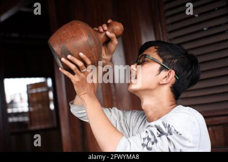 Porträt eines Mannes, der Wasser mit einem Kessel und Glas aus Ton trinkt. Traditionelle javanische Küchengeräte, die für warme Getränke verwendet werden. Indonesische rote Küche Stockfoto