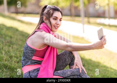 Junge kaukasische Frau auf Sportkleidung, die ein Selfie macht. Sie sitzt auf dem Gras eines grünen Parks Stockfoto